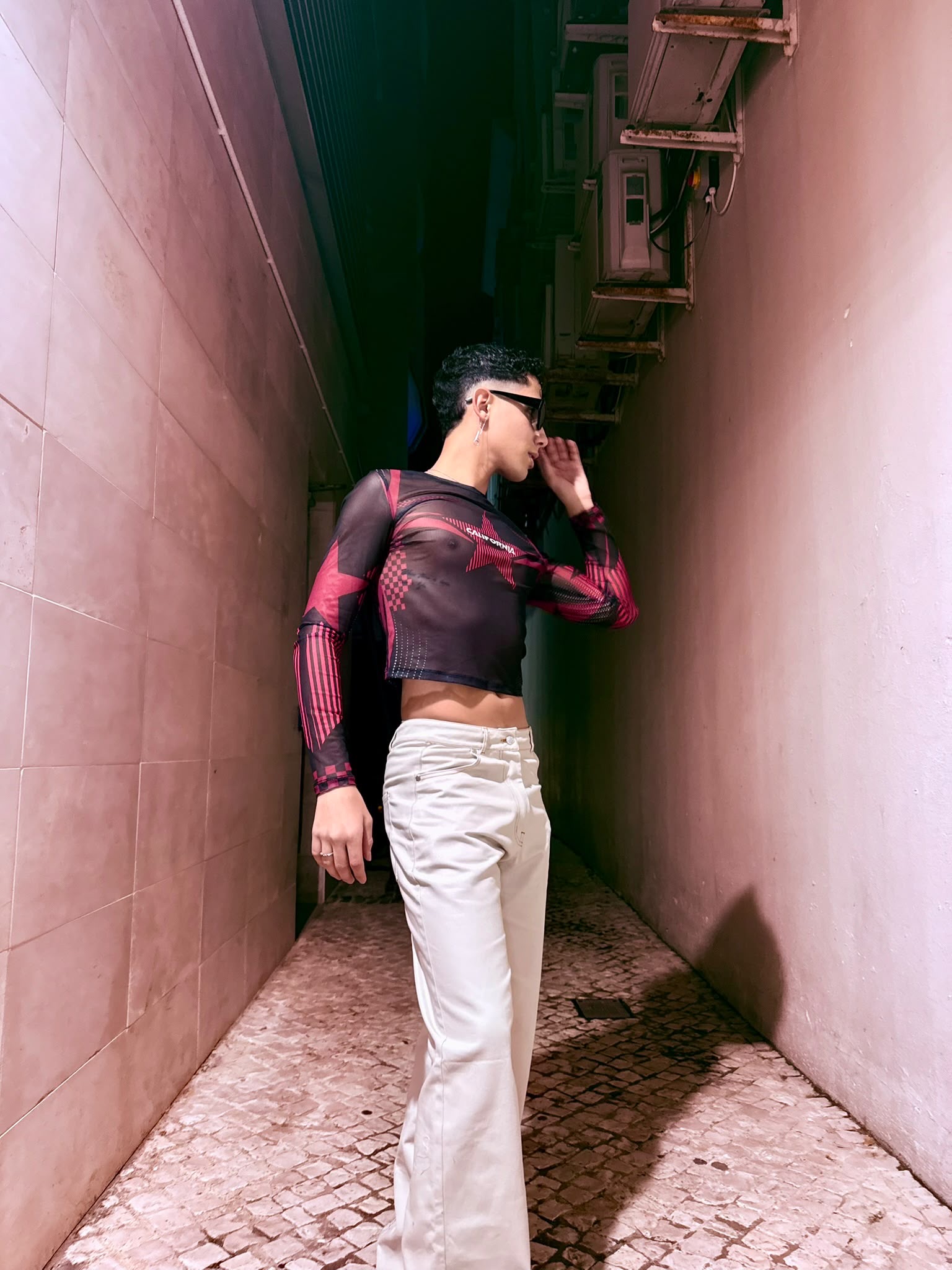 Karim Acolina wearing a sheer patterned top and wide white trousers in a pink alley.