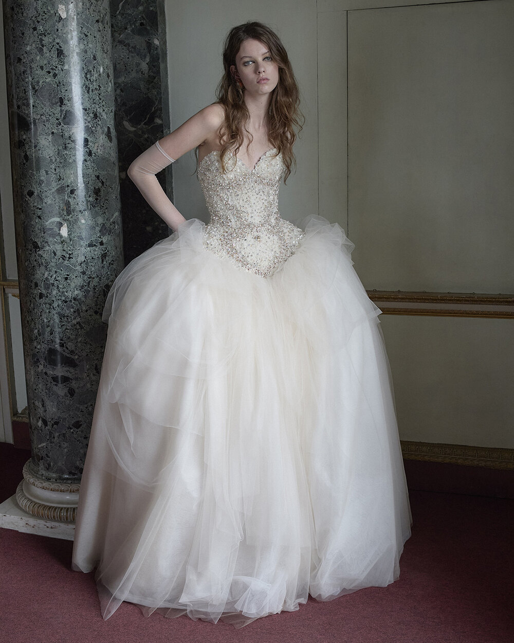 Mariposa bridal gown with a pointed pearl-, sequin- and crystal-embellished corset and a voluminous layered tulle skirt beside a green marble column.