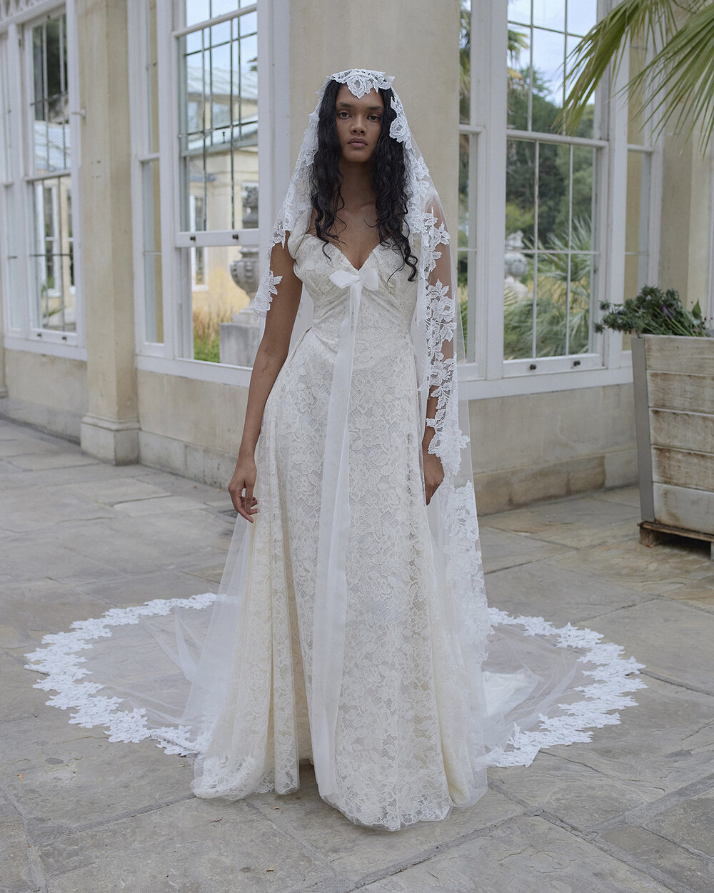 Reverie lace bridal gown with a sweetheart neckline, trailing organza bow and floral-edged veil in a conservatory.