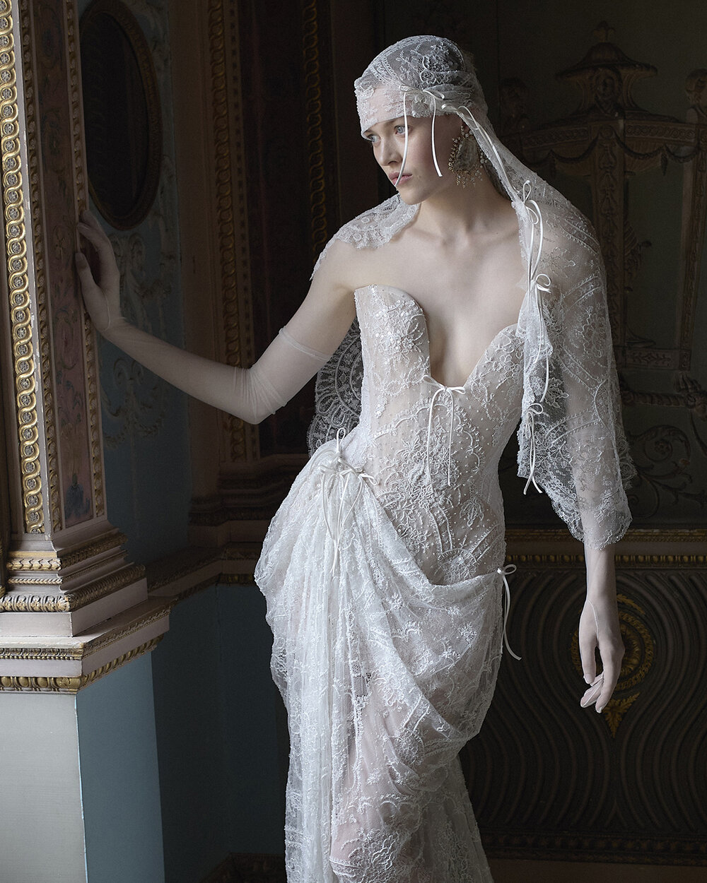 Seraphina lace bridal corset with a plunging neckline, delicate bows and lace draped over the model’s head beside a gilded mirror.